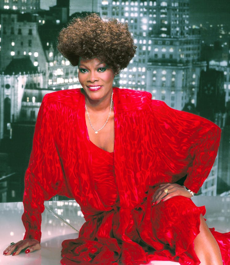 Dionne Warwick posing in a red dress with shoulder pads and her hand on her hip 