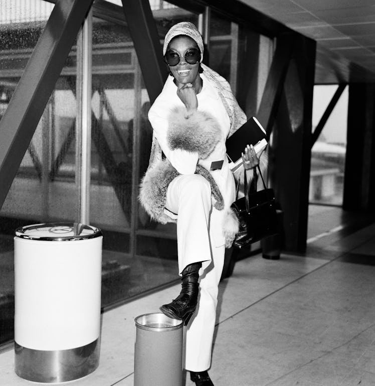 Dionne Warwick in white coat with fur sleeves, white pants and black boots, posing with her foot on ...