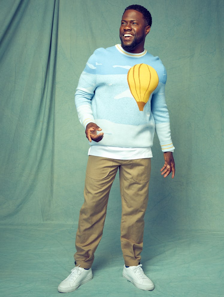 Kevin Hart stands in front of a green backdrop wearing a blue Victor Li sweater with a yellow hot ai...