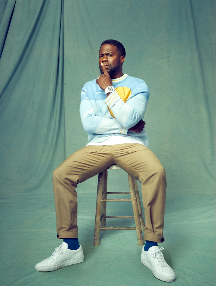 Kevin Hart sits on a stool in front of a green backdrop wearing a blue Victor Li sweater with a yell...