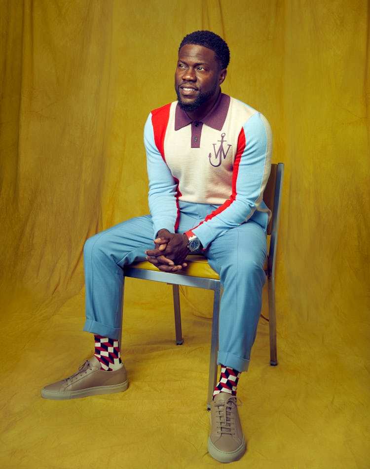 Kevin Hart wears a red, yellow, and blue JW Anderson sweater while sitting down in a chair for Rompe...