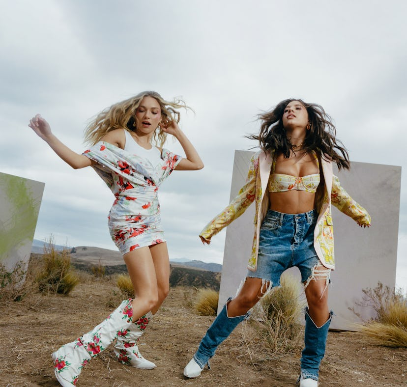 Two BFFs posing; a woman in a white floral dress an boors and a woman in a yellow blazer, bralette a...
