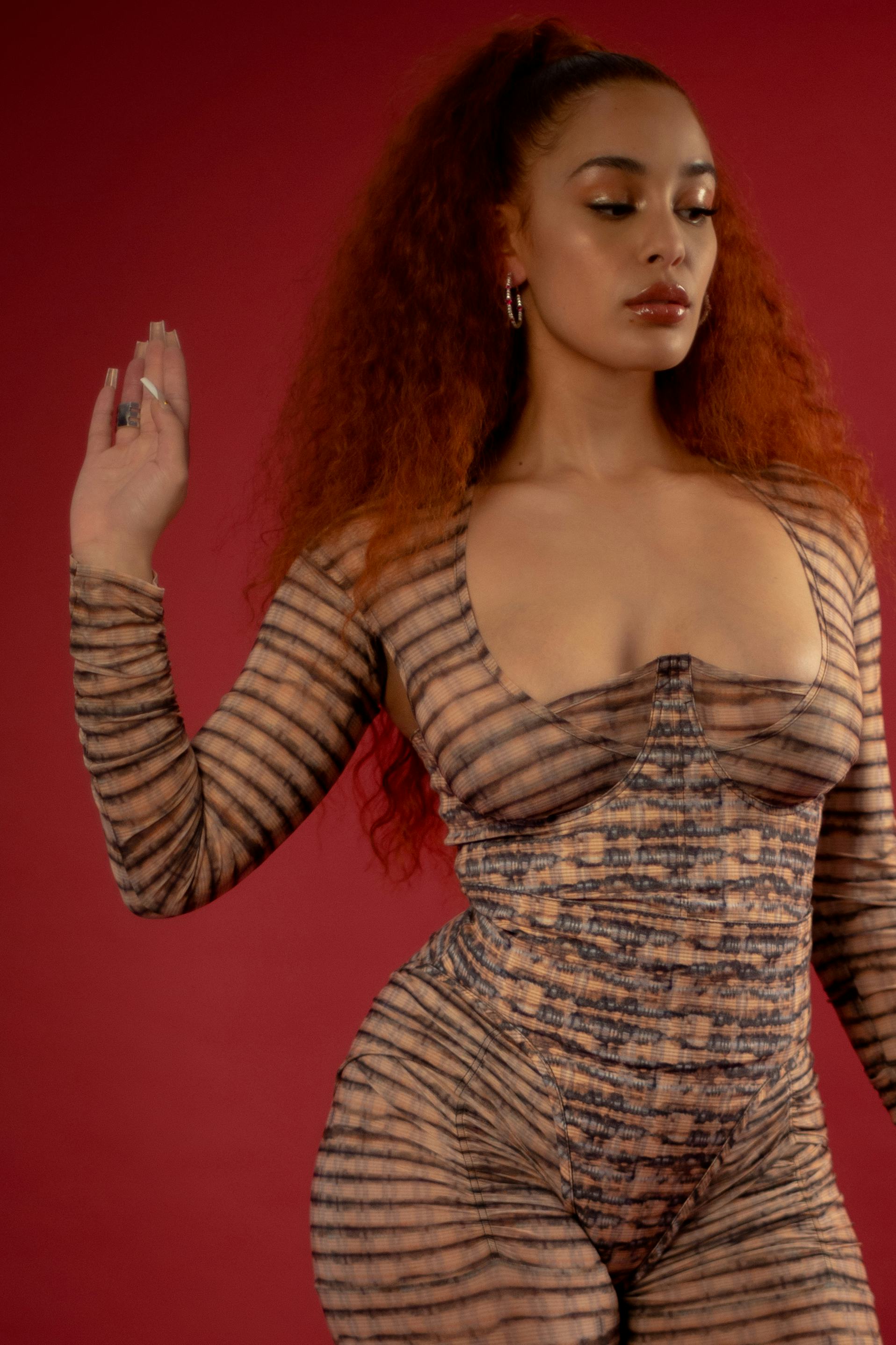 A photo of Jorja Smith. She's wearing a striped cat suit, and stands against a red background. Her e...