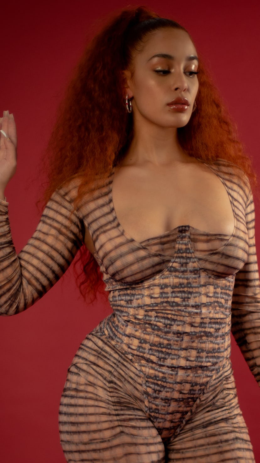 A photo of Jorja Smith. She's wearing a striped cat suit, and stands against a red background. Her e...