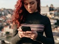 A red-haired woman in a black sweater with anxiety standing and using her smartphone and texting her...