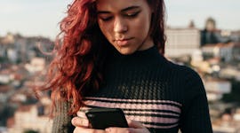 A red-haired woman in a black sweater with anxiety standing and using her smartphone and texting her...