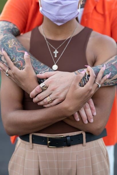 A man in an orange shirt, tattooed arms and two golden rings hugging a woman in a brown top from beh...