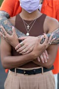 A man in an orange shirt, tattooed arms and two golden rings hugging a woman in a brown top from beh...