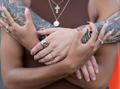 A man in an orange shirt, tattooed arms and two golden rings hugging a woman in a brown top from beh...