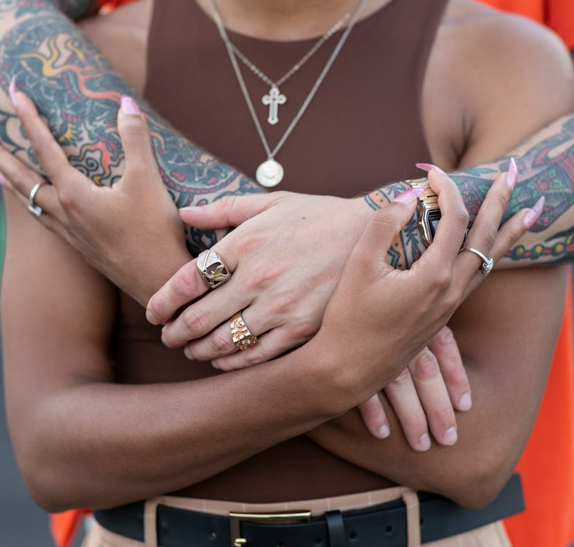A man in an orange shirt, tattooed arms and two golden rings hugging a woman in a brown top from beh...