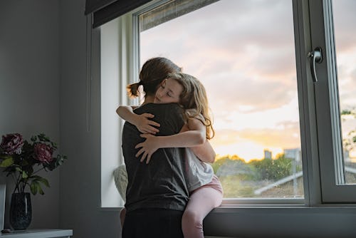 Young girl getting a big cuddle from her mother, in front of a large picture window.