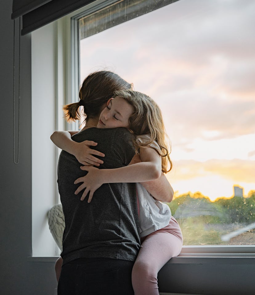 Young girl getting a big cuddle from her mother, in front of a large picture window.