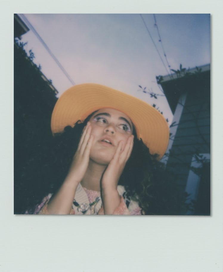 Remi Wolf with her hands on her face, wearing a yellow hat looking into the distance