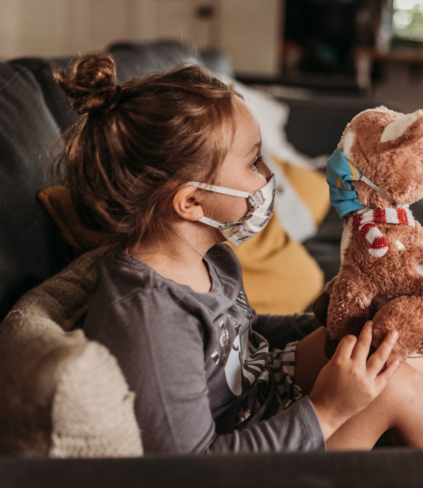 Side view of preschool age girl with mask on cuddling masked stuffed animal 