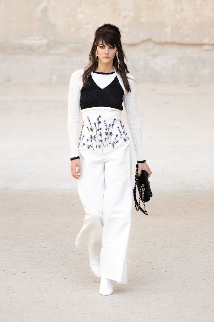 A model walking while wearing Chanel white pants and top, and a black crop top