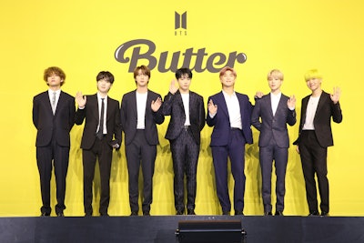 A photo of BTS. All 7 members are in black suits and standing against a yellow "Butter" press backgr...