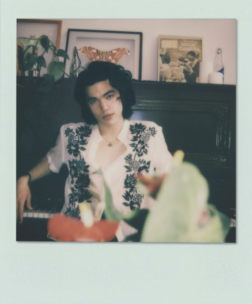 Conan Gray leaning against a piano in a white-black floral shirt with a blurred pastel color filter