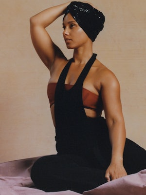 Alicia Keys sits on the ground wearing a black Hermes jumpsuit and black headpiece for Bustle's cove...