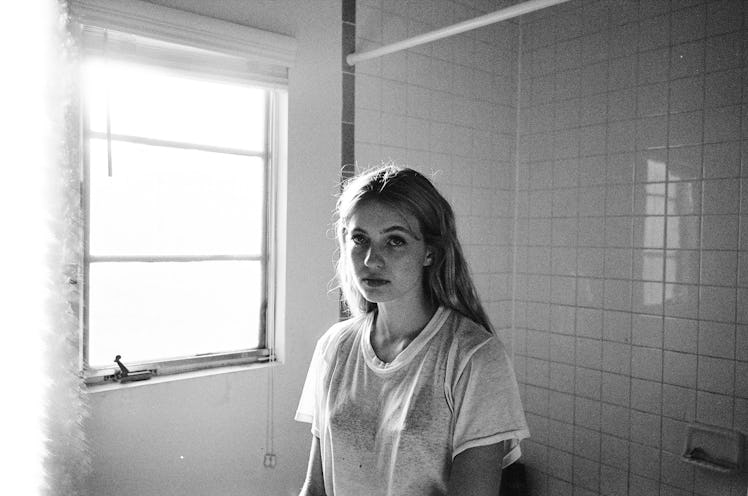 Black and white photo of Olivia Scott Welch in a white t-shirt, standing in a bathroom next to the w...
