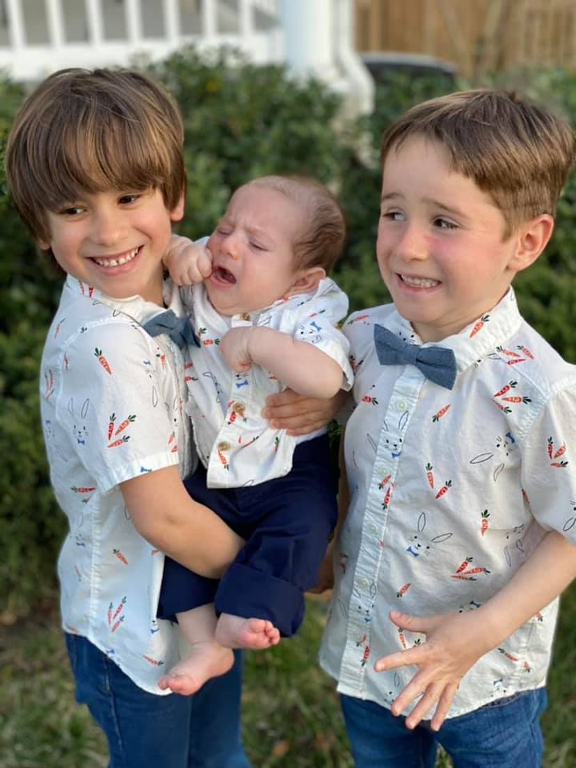 Twin boys hold their baby brother: one twin is beaming, the other looks concerned.