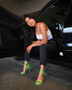 Janette Ok poses in white tanktop, black pants, and green heels.