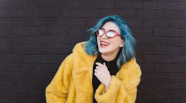 Smiling young woman with blue hair and yellow coat, having the best week of June 14, 2021, per her z...