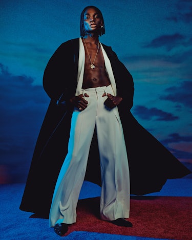Rickey Thompson stands against a dark sky background wearing a long, open black Chanel coat and whit...
