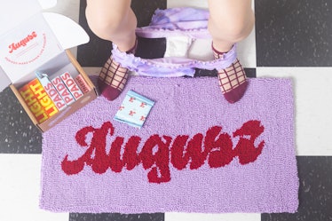 A girl sitting on a toilette seat while the box of August pads lies on the August rug on the floor n...