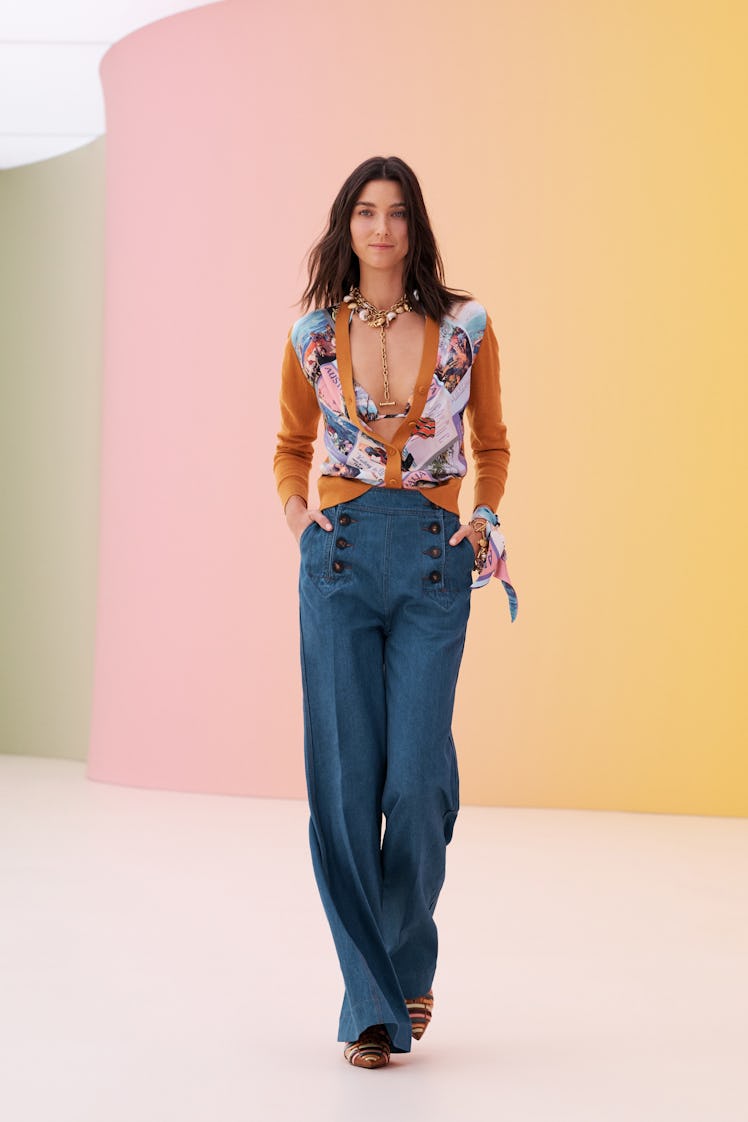 A woman posing while wearing blue pants and an orange and white blouse