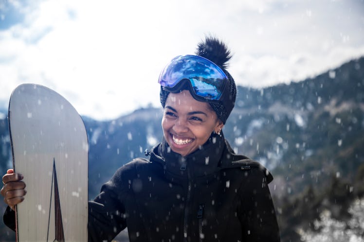 Smiling young woman in the snow, holding a snowboard ahead of posting on Instagram with a fun snowbo...