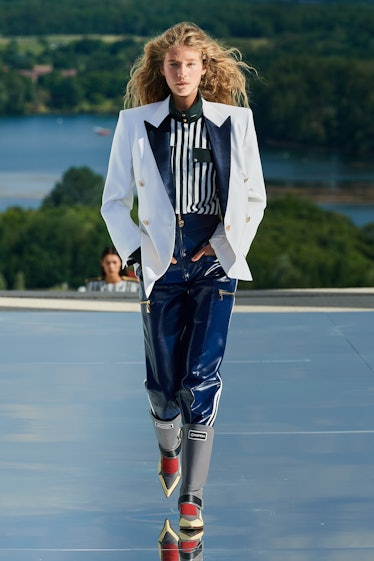 A female model walking while wearing a white blazer, a white and black striped shirt, and blue pants