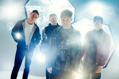Four members of Enter Shikari rock group standing in front of bright lights.