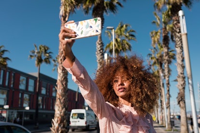 Young woman taking an elegant selfie with palm trees in the background, to post on Instagram with a ...