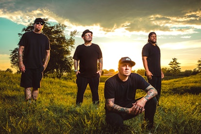The members of P.O.D posing in a field full of grass, with a sunset behind them. 