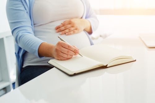 A pregnant woman sitting with one hand on her stomach and using her other hand to write in her pregn...