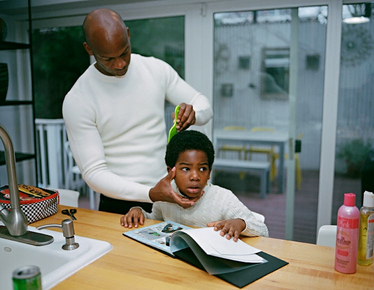 A man combing his son hair while he is reading a book on the kitchen counter