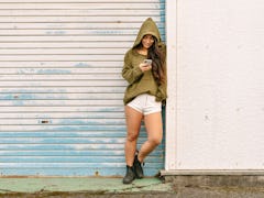 Young woman wearing a hoodie leaning against a garage, texting her friends funny private story names...