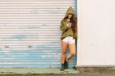 Young woman wearing a hoodie leaning against a garage, texting her friends funny private story names...