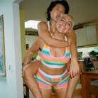 A woman with short blonde hair giving her queer partner a piggyback ride as they both wear rainbow o...