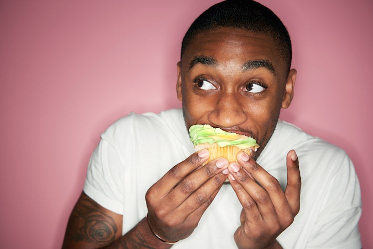 A man in a white shirt eating a cupcake in front of a pink wall