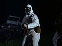 A man in a white hazmat suit standing in front of a truck holding a gun