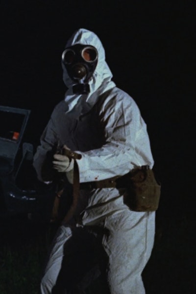 A man in a white hazmat suit standing in front of a truck holding a gun