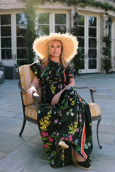 Kathy Hilton sits in a chair outside on a patio wearing a straw hat and a black floral dress.