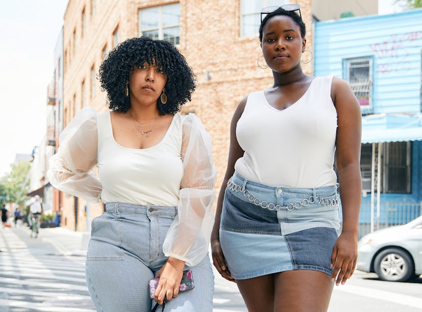 Two plus-size Black women standing on a street corner wearing plus-size white tops and denim bottoms...