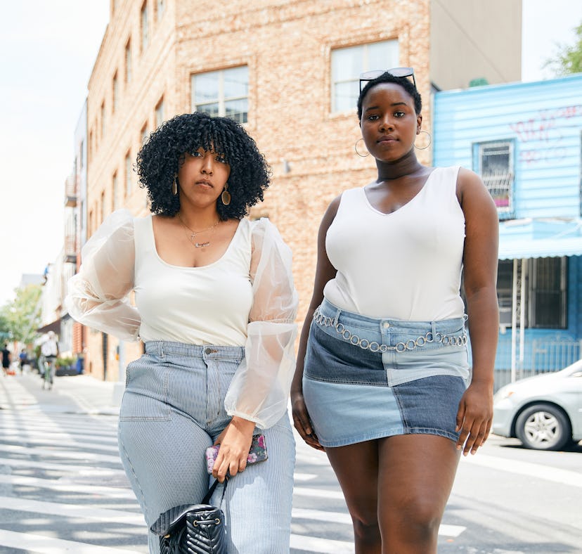 Two plus-size Black women standing on a street corner wearing plus-size white tops and denim bottoms...