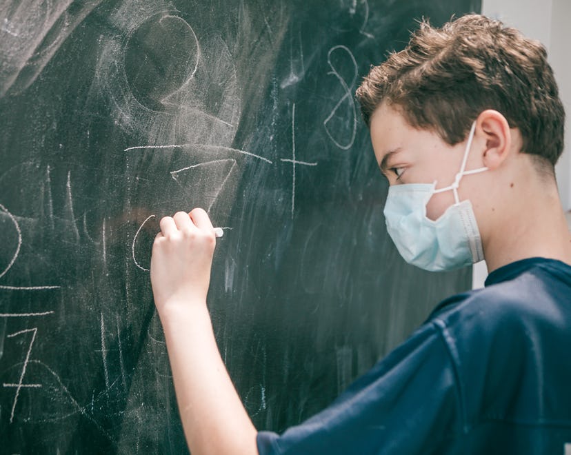 masked student in classroom working out math equations on chalkboard