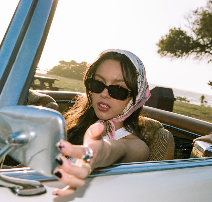 Olivia Rodrigo sitting in a car wearing items from her "deja vu" music video, which she is selling o...
