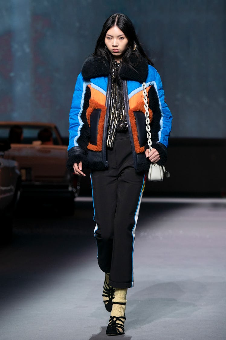 A female model walking the Coach Runway while wearing a blue, orange, and black jacket paired with b...