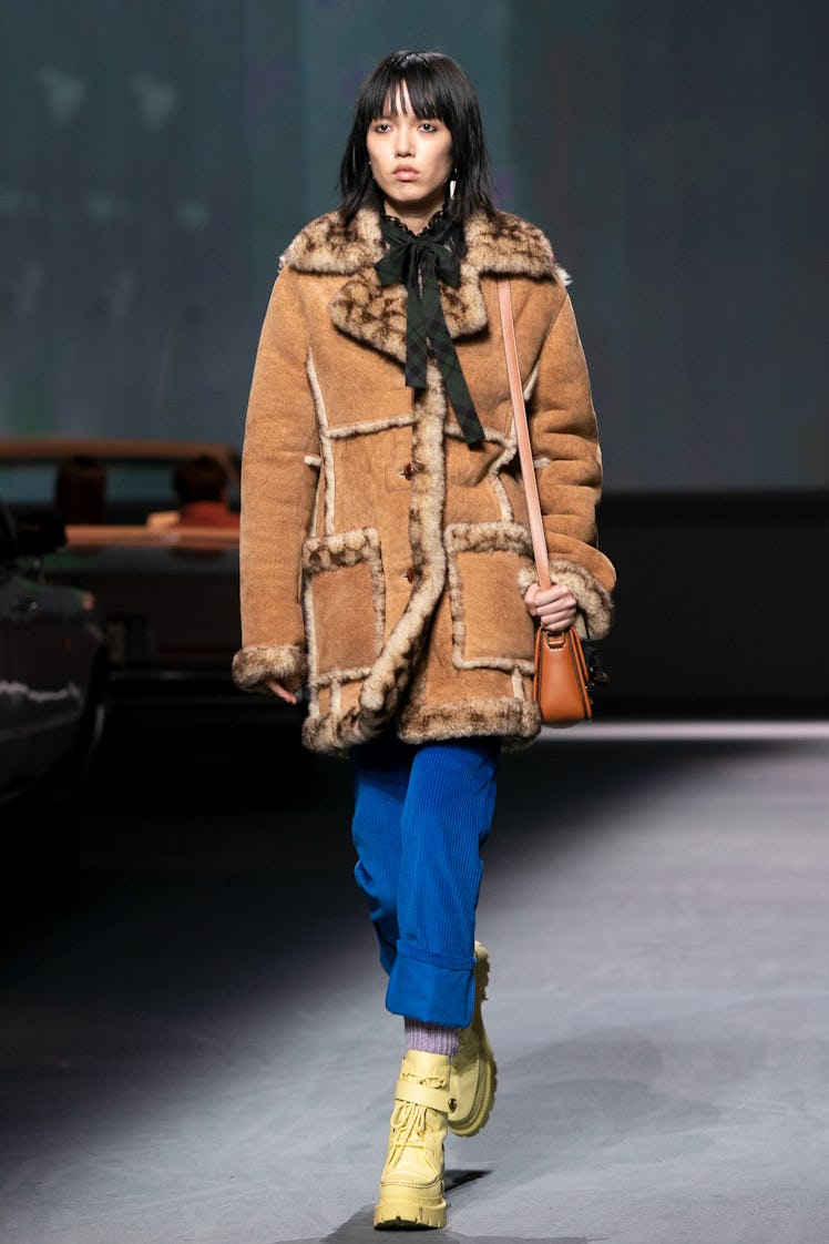 A female model walking the Coach Runway while wearing a brown jacket and blue pants