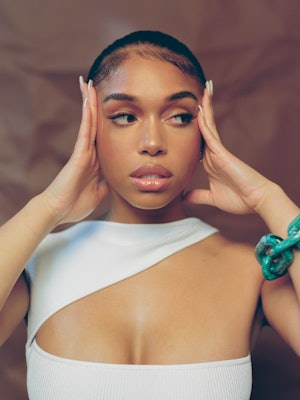 Model Lori Harvey poses in a white cut-out top and holds her hands to her head.